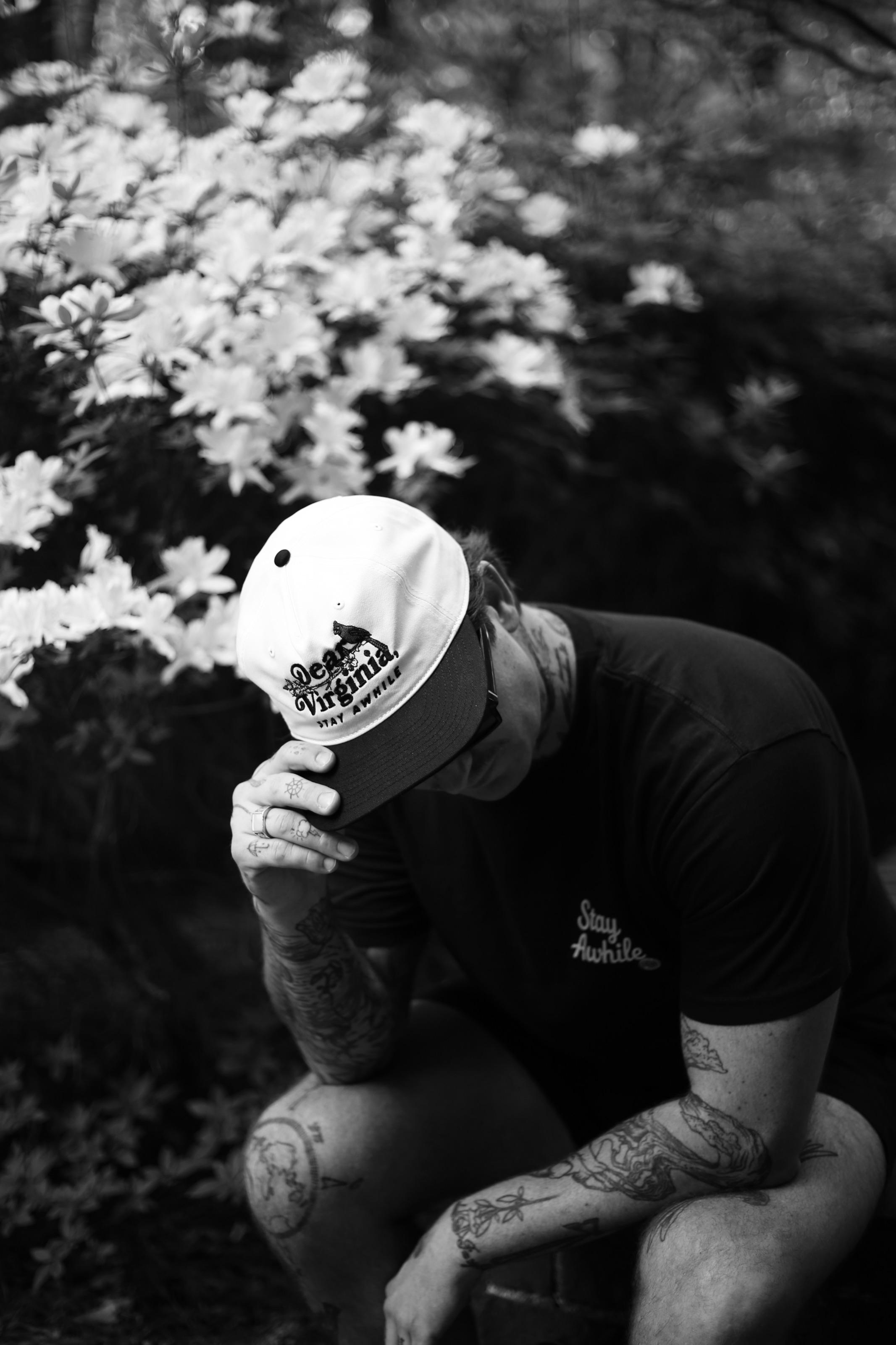 A person crouching in front of flowering azaleas, tipping down the brim of a Dear Virginia cap.