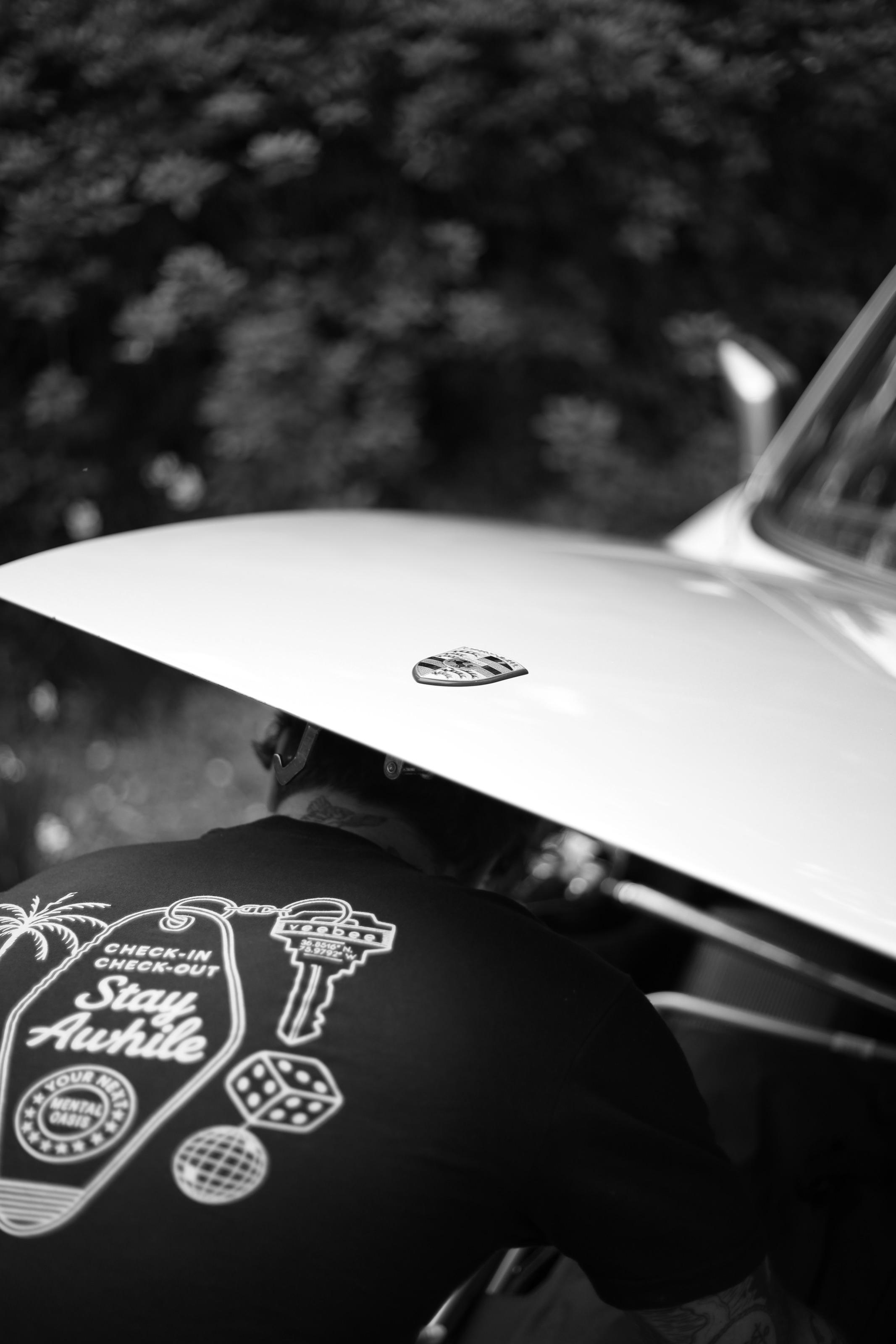 The back of a Stay Awhile t-shirt printed with a motel keytag, a room key, dice and a mirrorball, beneath the rear of a white Porsche.