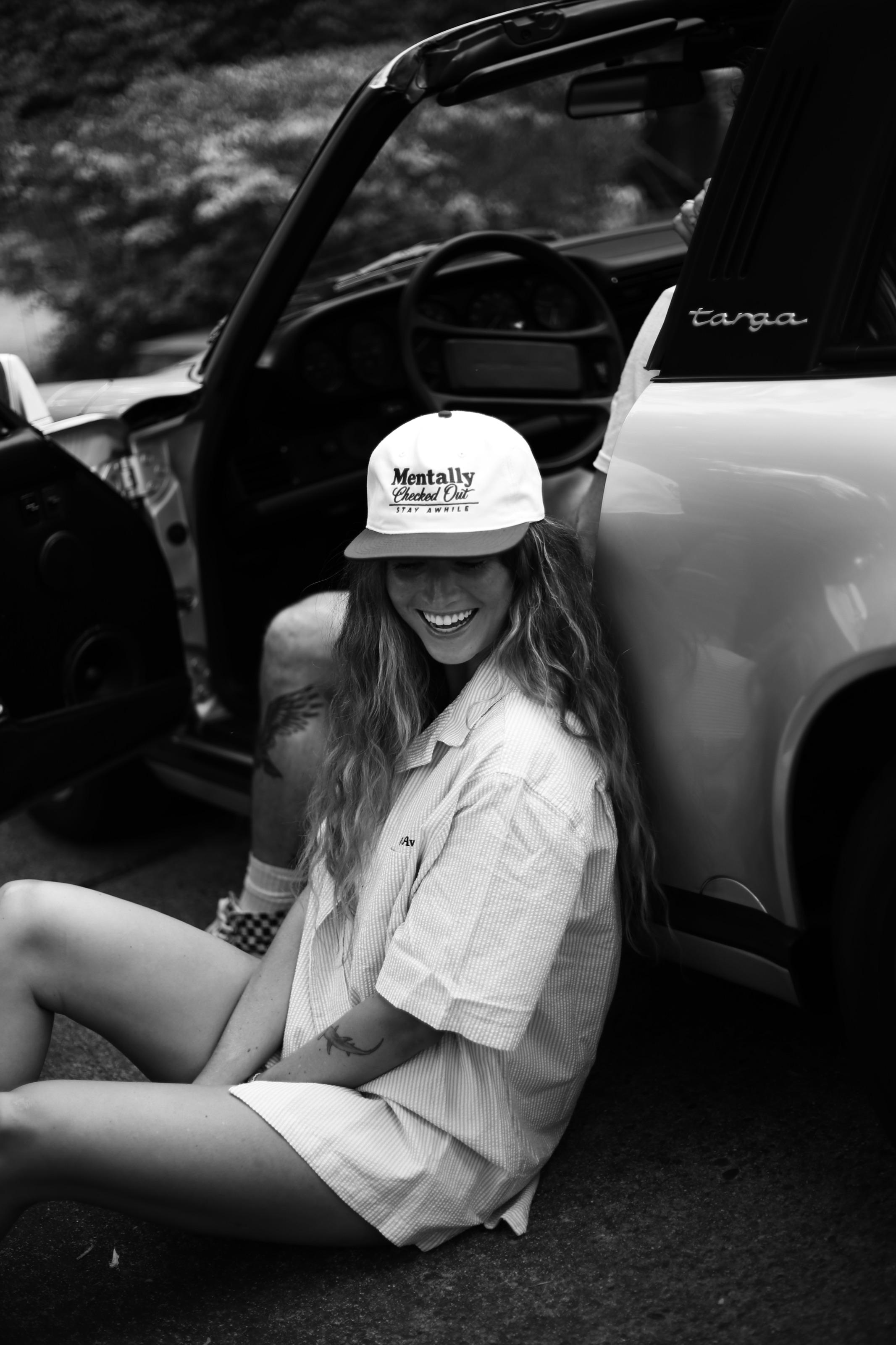 A person in a Mentally Checked Out cap sitting on the ground beside an open Porsche Targa door, laughing.